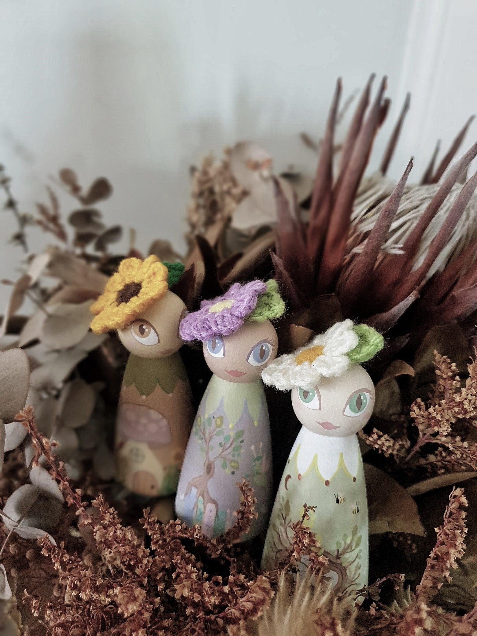 A set of three hand-painted wooden fairies, each with a different floral-themed design, positioned among dried flowers.