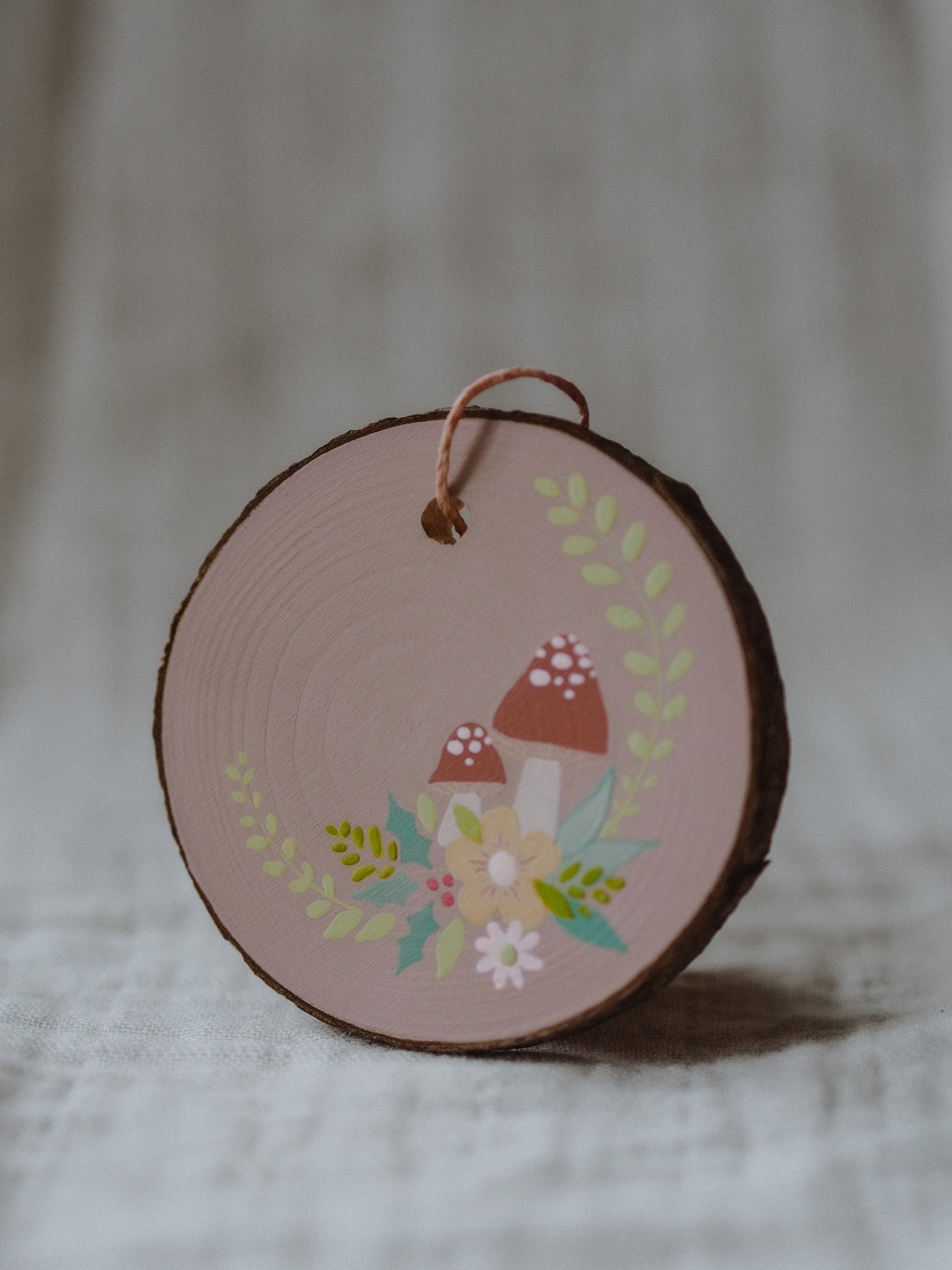 A round, hand-painted wood slice ornament with a Christmas design, featuring a neutral color palette and decorative elements such as a house, tree, and flowers.