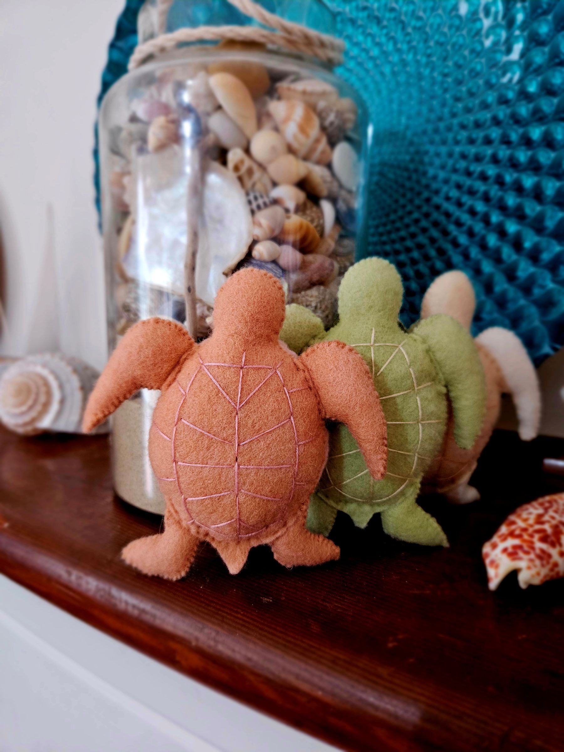 Two felt turtles in front of a jar filled with seashells on a wooden surface.