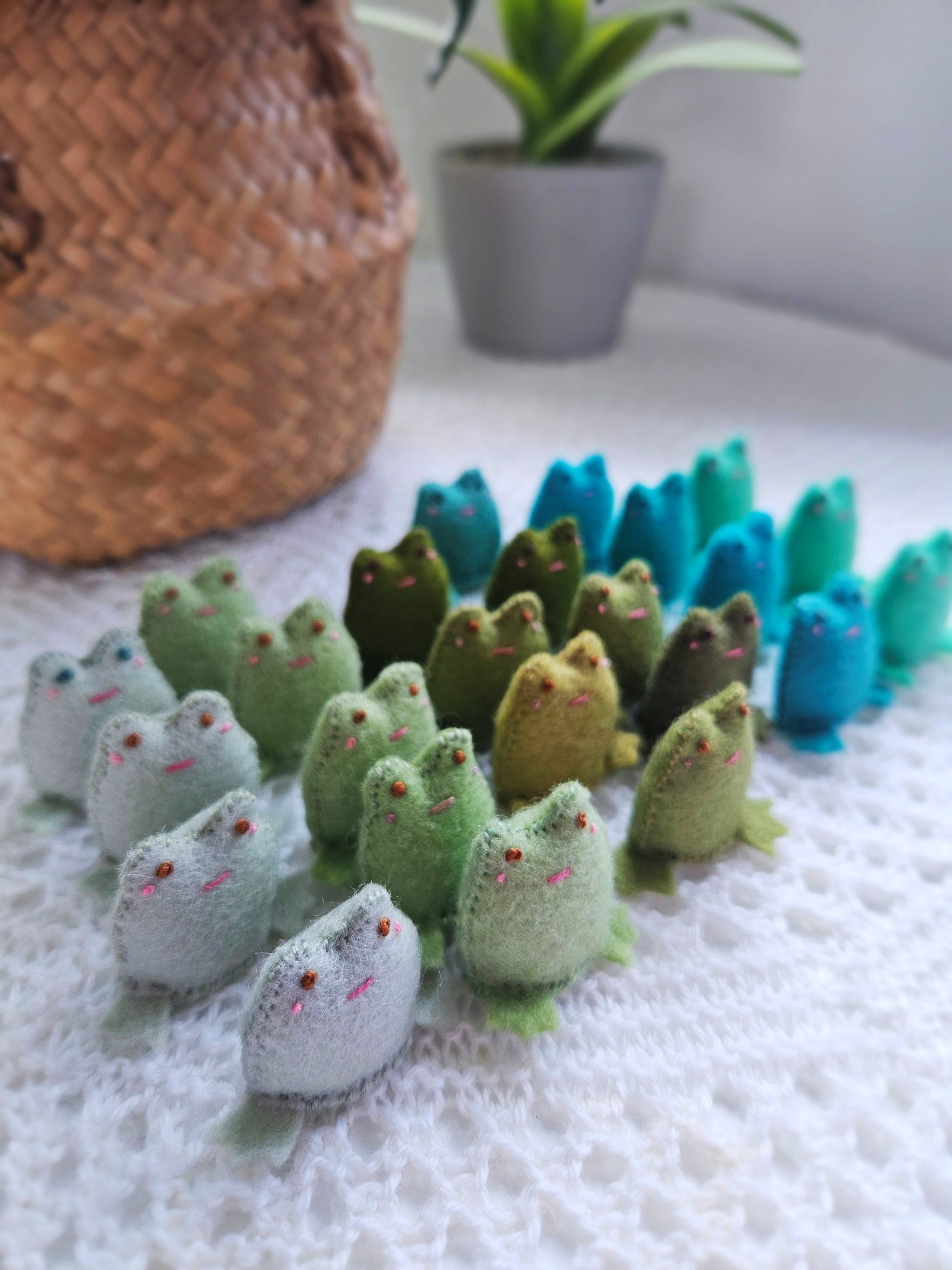 Collection of small wool felt frogs on a textured surface with a plant in the background.
